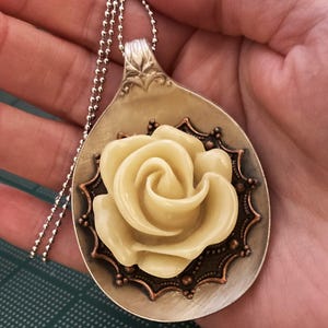 May include: A silver spoon necklace with a cream-colored rose and copper accents. The rose is centered on the spoon and surrounded by a decorative copper frame.