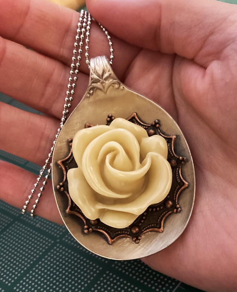 May include: A silver spoon necklace with a cream-colored rose and copper accents. The rose is centered on the spoon and surrounded by a decorative copper frame.