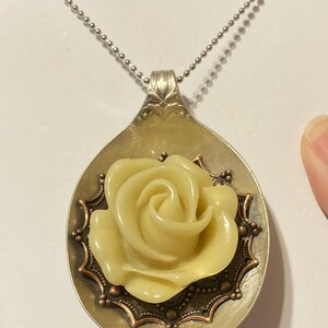 May include: A silver spoon necklace with a yellow rose charm. The rose is surrounded by a copper-colored filigree design.