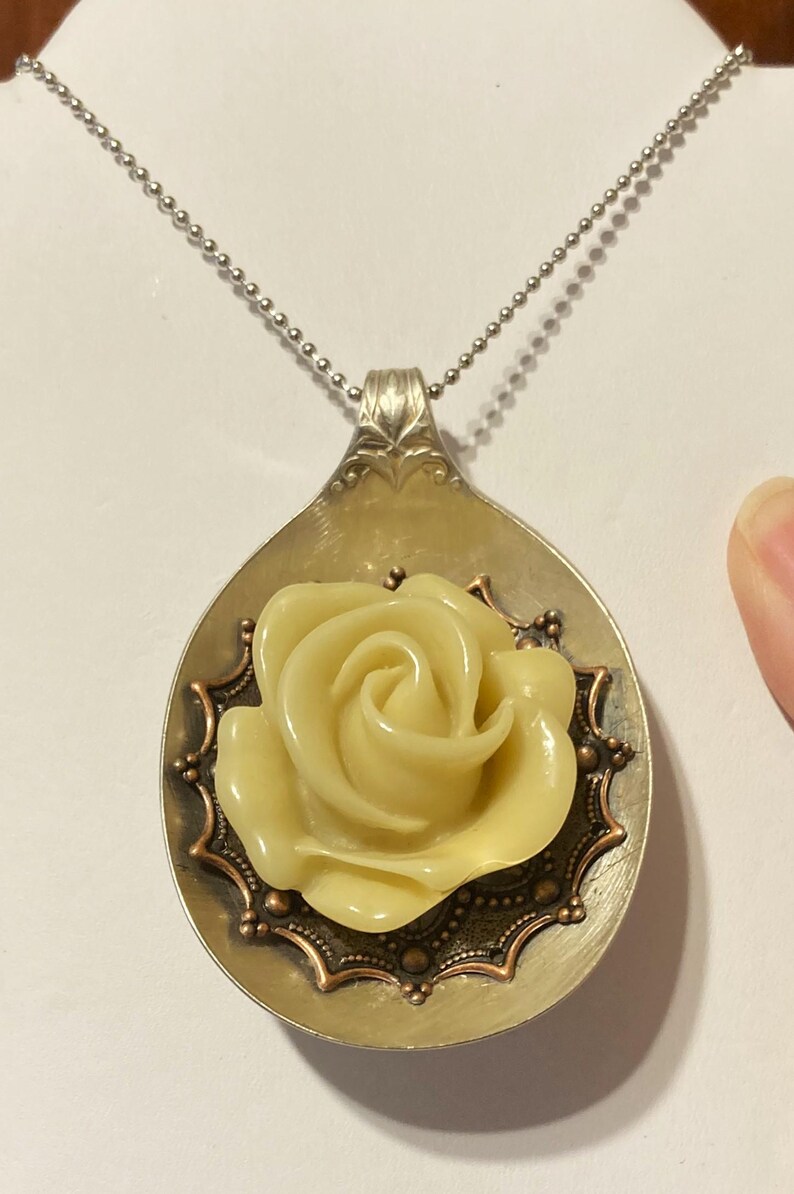 May include: A silver spoon necklace with a yellow rose charm. The rose is surrounded by a copper-colored filigree design.