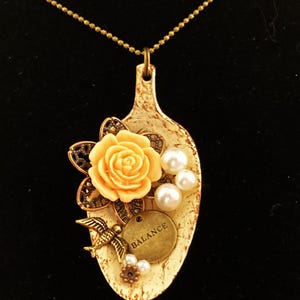 May include: A white spoon pendant necklace with a peach-colored rose, pearl accents, a small bird charm, and a gold charm that says "BALANCE".