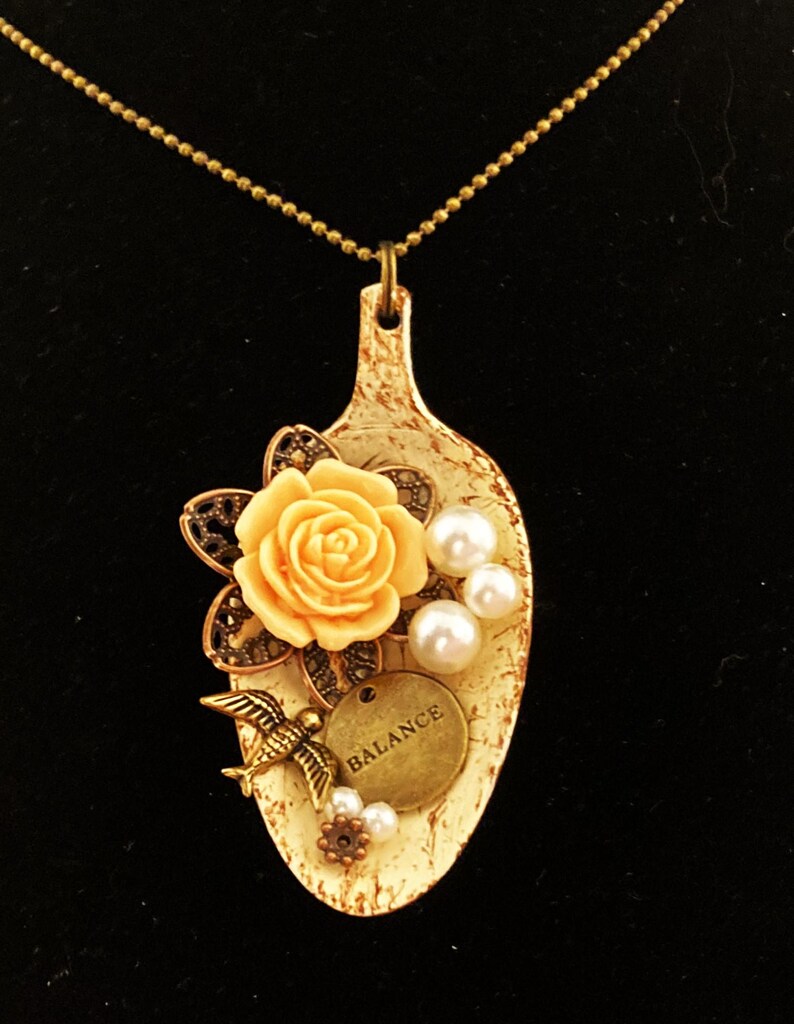 May include: A white spoon pendant necklace with a peach-colored rose, pearl accents, a small bird charm, and a gold charm that says "BALANCE".