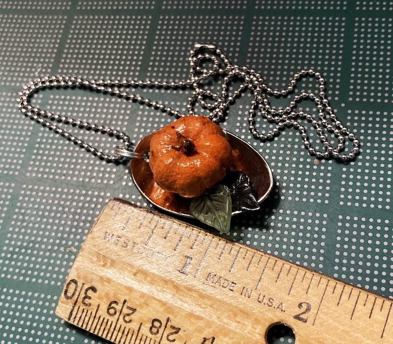 May include: A silver chain necklace with a pendant featuring a small orange pumpkin on a spoon. The spoon is brown and has a green leaf attached to it. The necklace is on a green background with white dots.