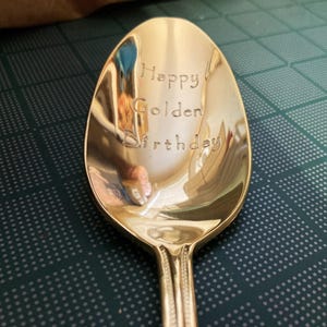 May include: A gold-colored spoon with the words "Happy Golden Birthday" engraved on the bowl. The spoon has a decorative handle and reflects the surrounding environment. The spoon is on a dark green surface.