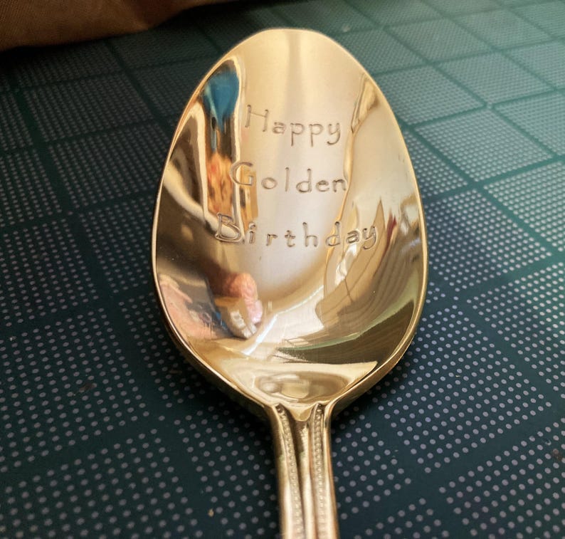 May include: A gold-colored spoon with the words "Happy Golden Birthday" engraved on the bowl. The spoon has a decorative handle and reflects the surrounding environment. The spoon is on a dark green surface.