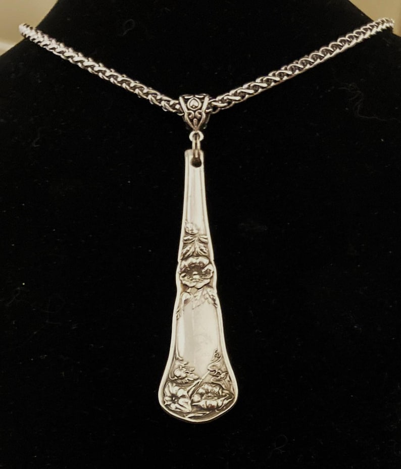 May include: A silver necklace with a long, ornate pendant made from a vintage spoon. The pendant features a floral design.