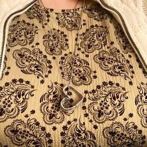 May include: A silver heart-shaped pendant necklace on a silver chain. The necklace is worn over a beige top with a brown paisley pattern.