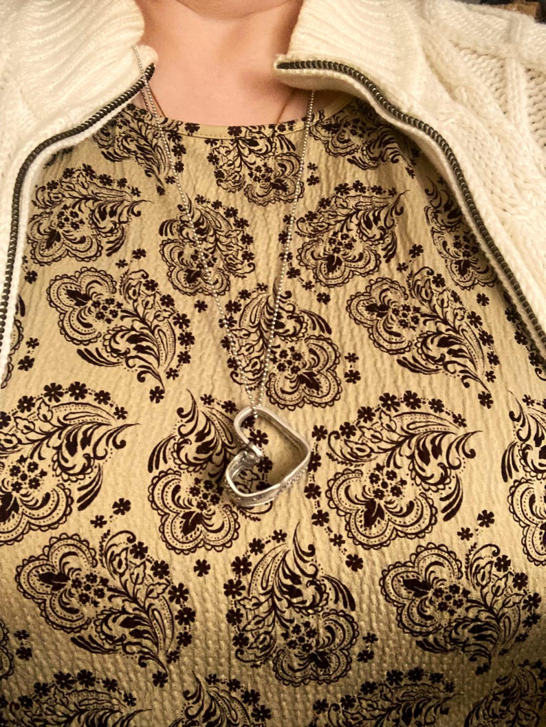 May include: A silver heart-shaped pendant necklace on a silver chain. The necklace is worn over a beige top with a brown paisley pattern.