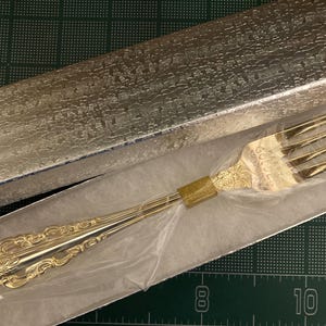 May include: A gold-colored fork with ornate detailing, presented in a silver textured box. The fork has four tines and a decorative handle. The box is open, revealing the fork inside. The fork is likely a piece of silverware.