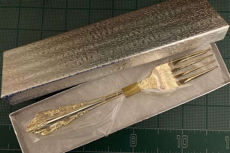 May include: A gold-colored fork with ornate detailing, presented in a silver textured box. The fork has four tines and a decorative handle. The box is open, revealing the fork inside. The fork is likely a piece of silverware.