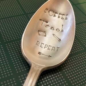 May include: A silver spoon with the words "Coffee Teach Repeat" engraved on it. The spoon also has small illustrations of a coffee cup, a book, a pencil, and an arrow.
