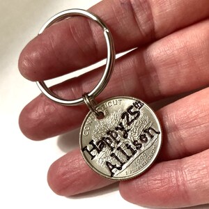 May include: A silver keychain with a quarter coin pendant. The coin is engraved with "Happy 25th Allison" and the year 1999. The keychain is held by a person.