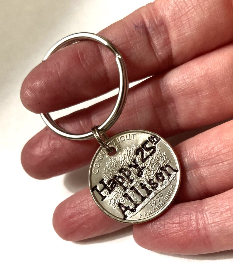 May include: A silver keychain with a quarter coin pendant. The coin is engraved with "Happy 25th Allison" and the year 1999. The keychain is held by a person.