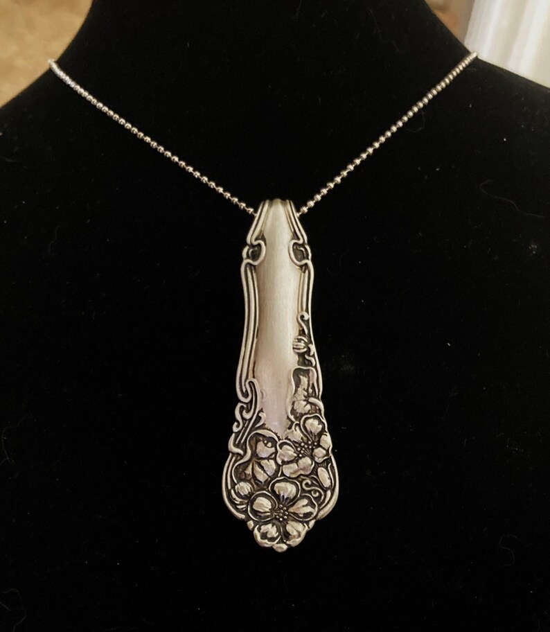 May include: A silver spoon necklace with a floral design. The spoon is attached to a silver chain.