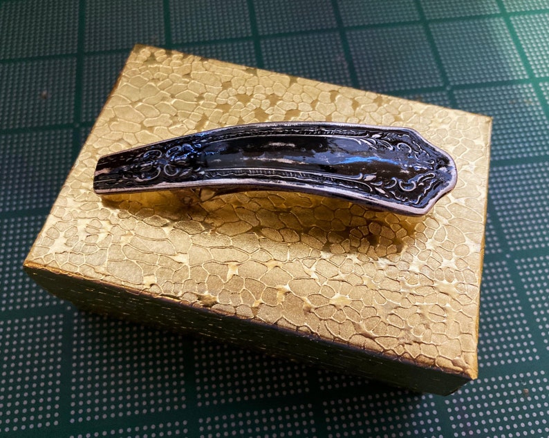 May include: A silver-toned hair clip with an ornate design. The clip is black and has a decorative pattern. The clip is sitting on a gold-colored surface.