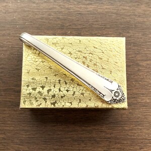 May include: A silver-colored spoon with a floral design on the handle rests on a gold-colored, textured box. The spoon is angled across the box, showcasing its intricate details and polished finish. The background is a dark wood surface.