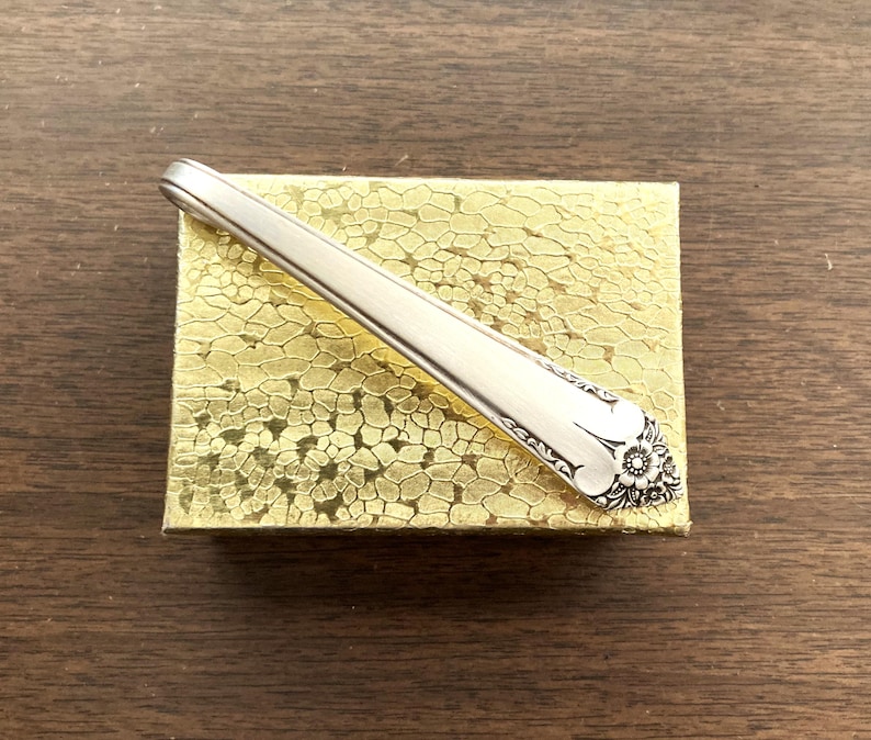 May include: A silver-colored spoon with a floral design on the handle rests on a gold-colored, textured box. The spoon is angled across the box, showcasing its intricate details and polished finish. The background is a dark wood surface.