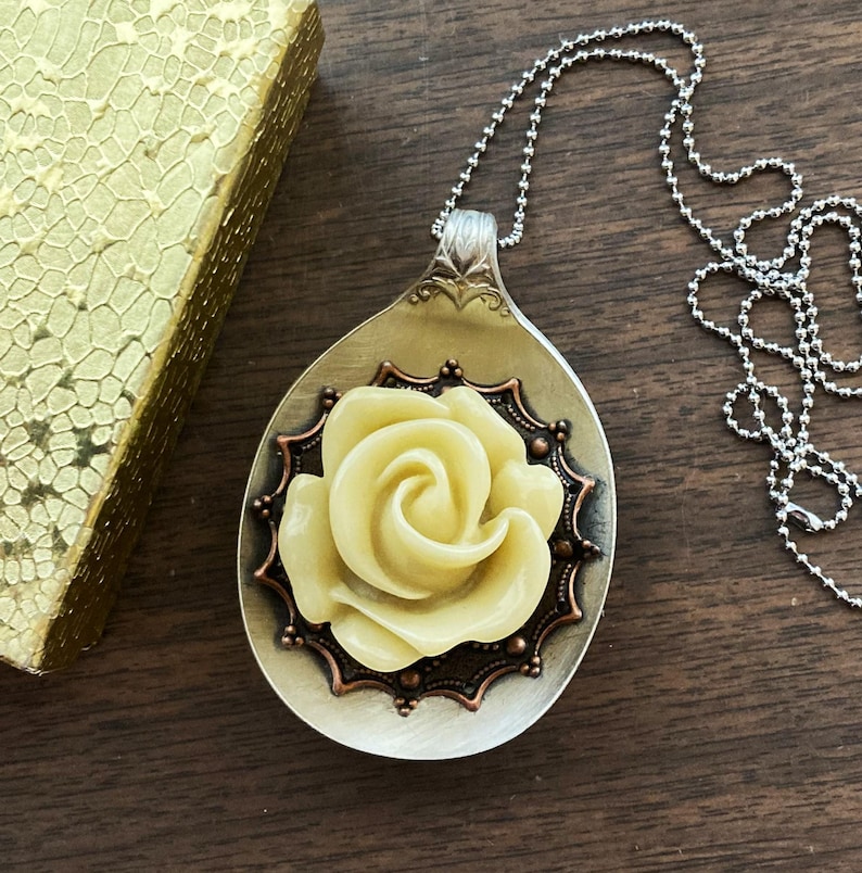May include: A silver spoon necklace with a yellow rose charm. The rose is set in a copper-colored frame with a decorative design.