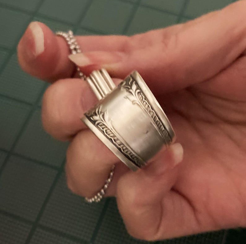 May include: A silver spoon ring with a decorative design. The ring is made from a repurposed spoon.