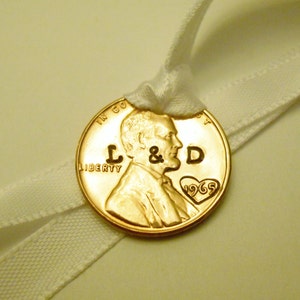 K&ouml;nnte beinhalten: Ein goldfarbener US-Penny mit den Worten "In God We Trust" und "Liberty", die darauf eingraviert sind. Die Initialen "L & D" sind auf der M&uuml;nze eingraviert, zusammen mit einem Herzen, das das Jahr "1965" enth&auml;lt. Die M&uuml;nze ist mit einem wei&szlig;en Band gebunden.