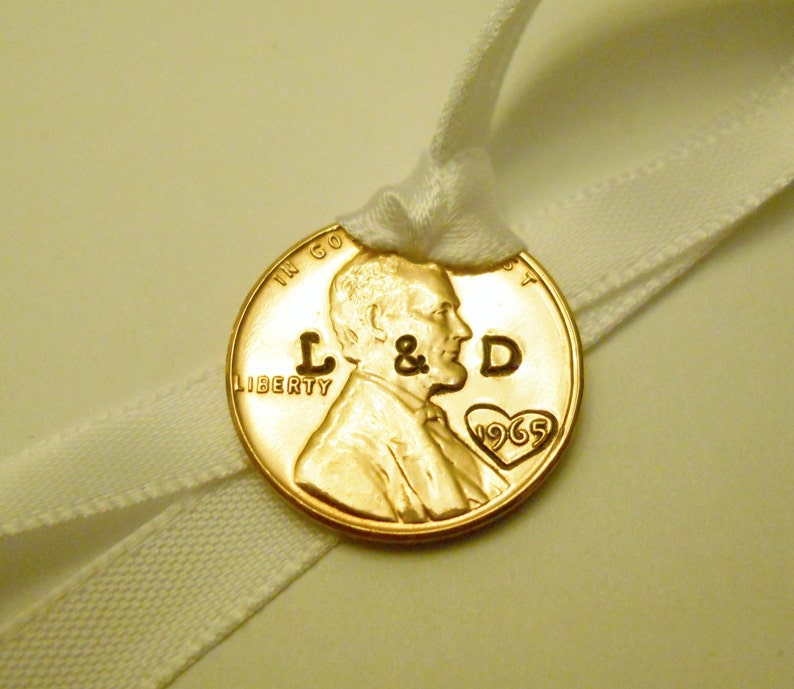 K&ouml;nnte beinhalten: Ein goldfarbener US-Penny mit den Worten "In God We Trust" und "Liberty", die darauf eingraviert sind. Die Initialen "L & D" sind auf der M&uuml;nze eingraviert, zusammen mit einem Herzen, das das Jahr "1965" enth&auml;lt. Die M&uuml;nze ist mit einem wei&szlig;en Band gebunden.