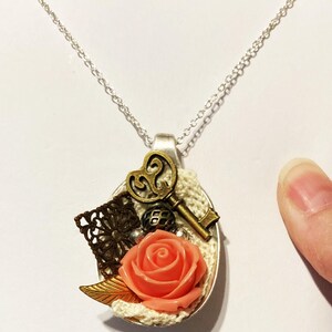 May include: A silver chain necklace with a spoon pendant. The spoon is decorated with a coral-colored rose, a key, a small metal ball, and a piece of lace.