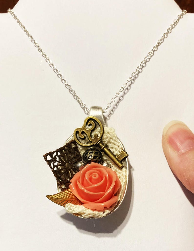 May include: A silver chain necklace with a spoon pendant. The spoon is decorated with a coral-colored rose, a key, a small metal ball, and a piece of lace.