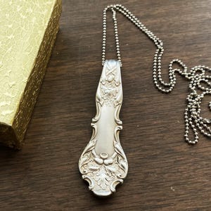 Puede incluir: Un collar de plata con un colgante de cubiertos vintage. El colgante es una cuchara larga y adornada con un dise&ntilde;o floral.