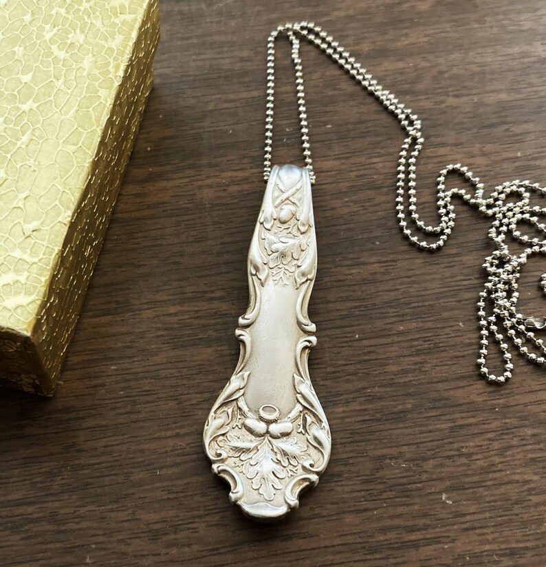 Puede incluir: Un collar de plata con un colgante de cubiertos vintage. El colgante es una cuchara larga y adornada con un dise&ntilde;o floral.