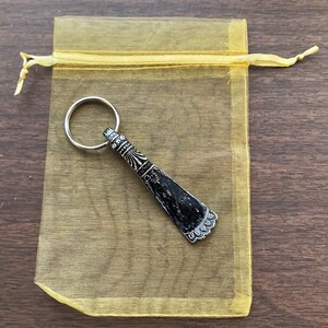 May include: A silver shoehorn keychain with an ornate design and a black handle. The keychain is in a yellow organza bag.