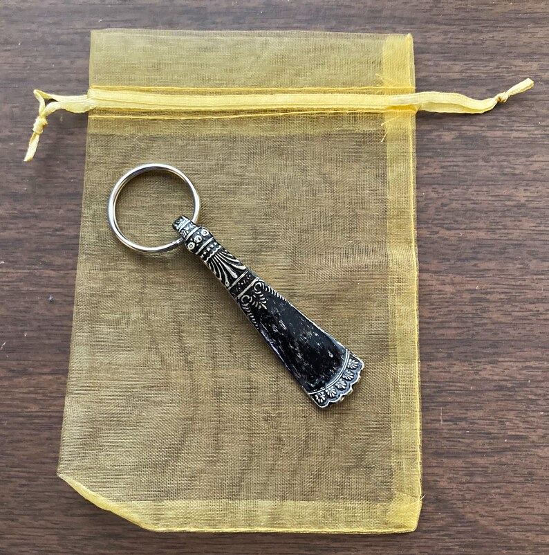 May include: A silver shoehorn keychain with an ornate design and a black handle. The keychain is in a yellow organza bag.