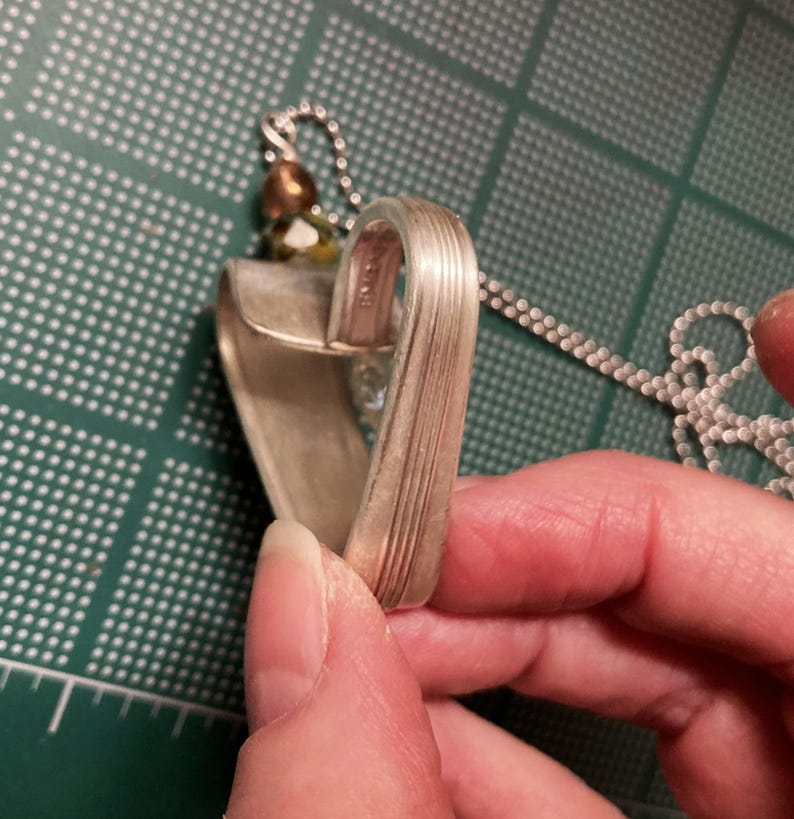 May include: A silver heart-shaped pendant necklace made from a repurposed spoon. The pendant has a textured surface and is attached to a silver chain.