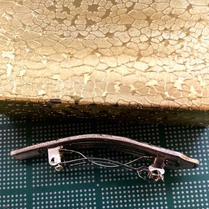 May include: A silver hair clip with a curved, rectangular shape and a spring mechanism. The clip is on a green surface with a pattern of small white dots.