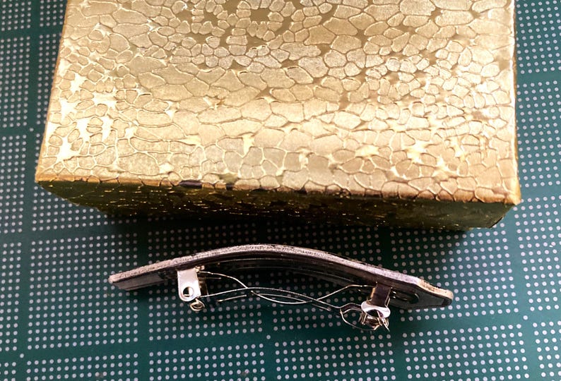 May include: A silver hair clip with a curved, rectangular shape and a spring mechanism. The clip is on a green surface with a pattern of small white dots.