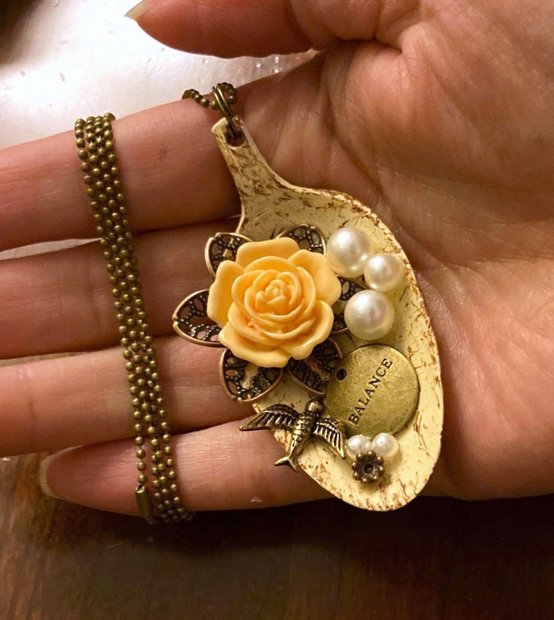 May include: A necklace with a spoon pendant featuring a yellow rose, a bird, pearls, and a charm that says "Balance".
