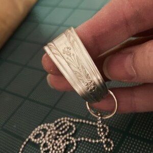 May include: A silver necklace with a pendant made from a repurposed spoon. The spoon has an ornate floral design and is attached to a silver chain.