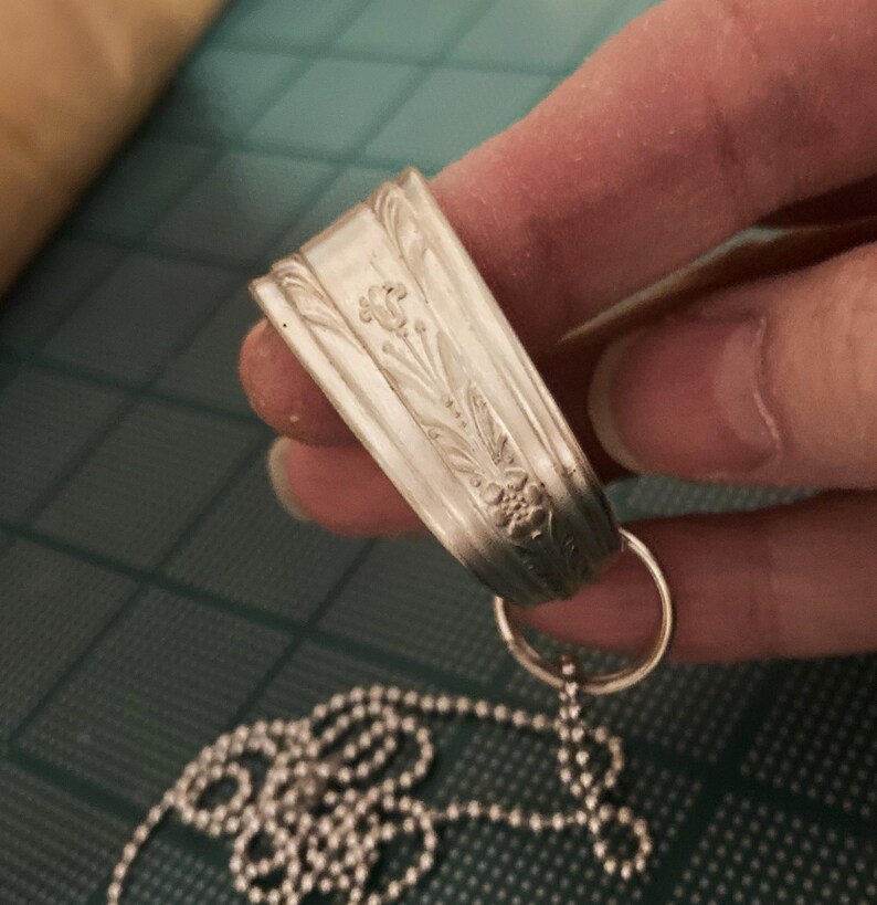 May include: A silver necklace with a pendant made from a repurposed spoon. The spoon has an ornate floral design and is attached to a silver chain.