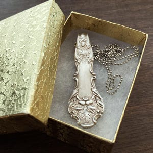 Puede incluir: Un collar de cuchara de plata con un dise&ntilde;o ornamentado, que presenta un patr&oacute;n floral y un peque&ntilde;o amuleto de bellota. El collar se muestra en una caja de regalo dorada con papel de seda blanco.