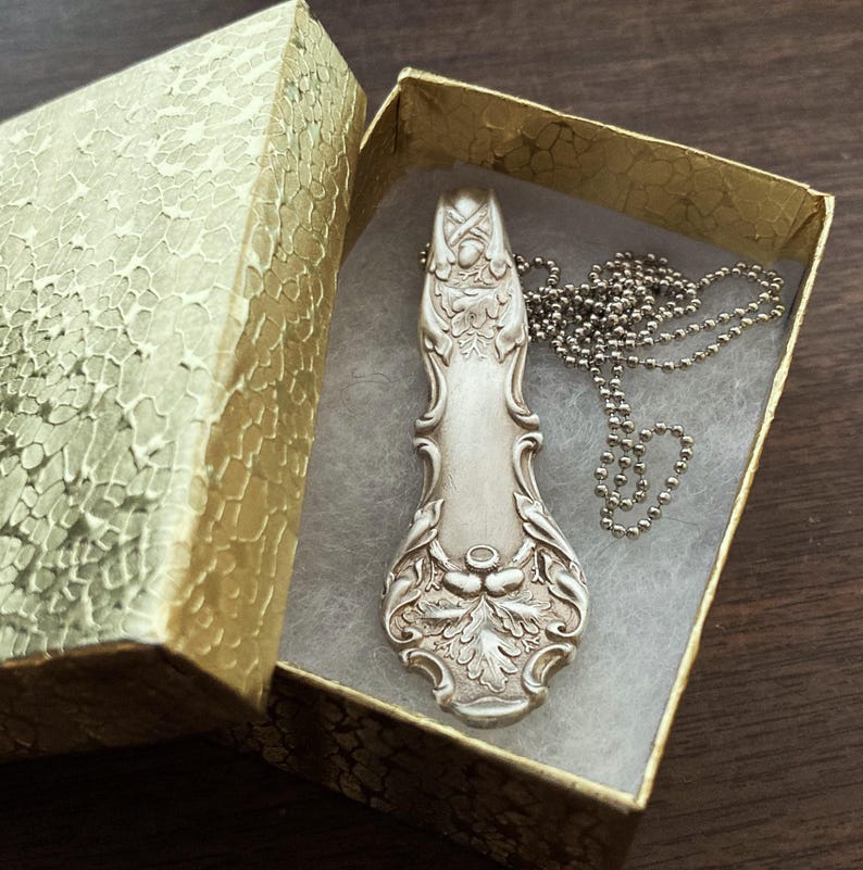 Puede incluir: Un collar de cuchara de plata con un dise&ntilde;o ornamentado, que presenta un patr&oacute;n floral y un peque&ntilde;o amuleto de bellota. El collar se muestra en una caja de regalo dorada con papel de seda blanco.