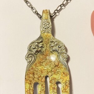 May include: A silver fork necklace with a yellow and brown marbled design. The fork has a floral design on the handle and is attached to a silver chain.
