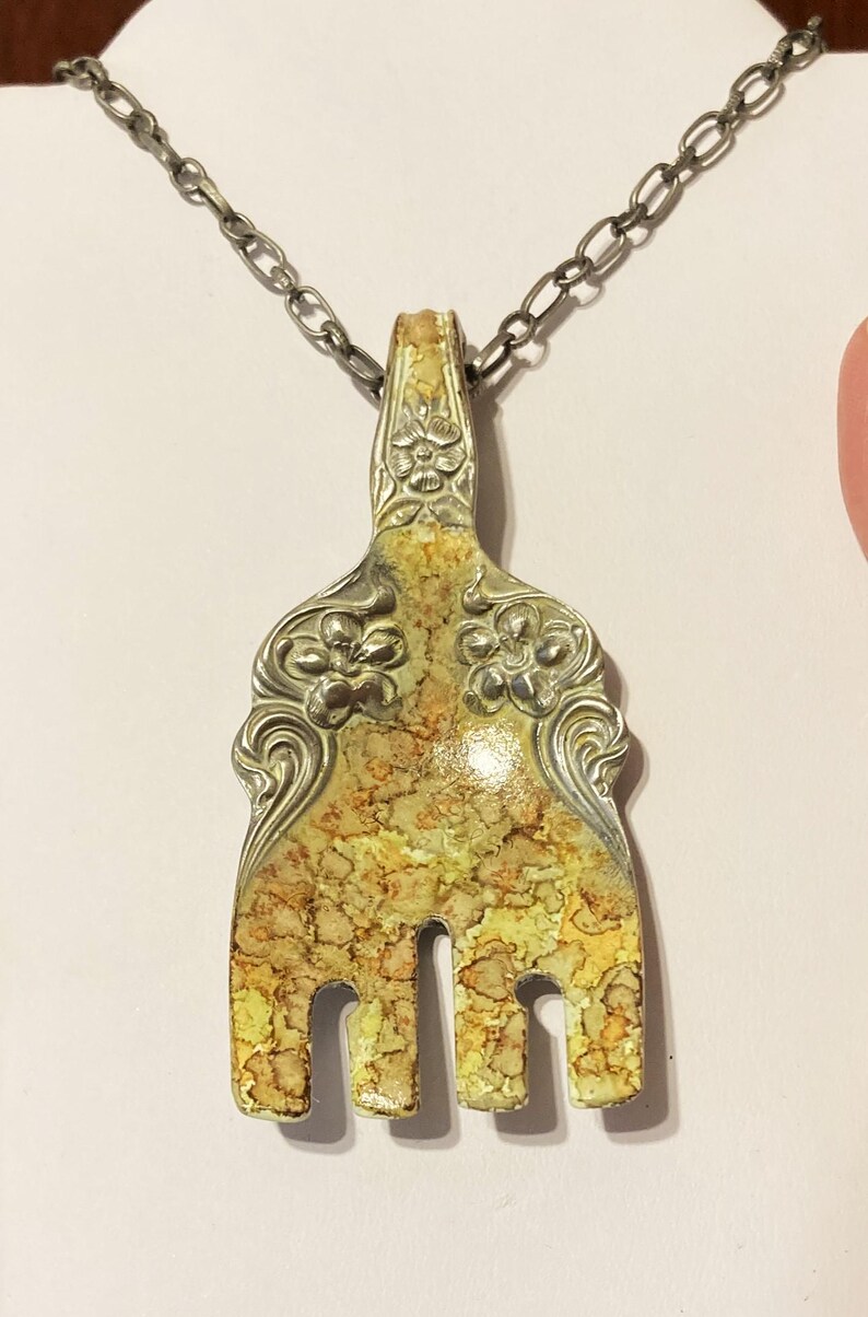 May include: A silver fork necklace with a yellow and brown marbled design. The fork has a floral design on the handle and is attached to a silver chain.