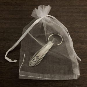 May include: A silver spoon keychain with a decorative design, packaged in a white organza bag.