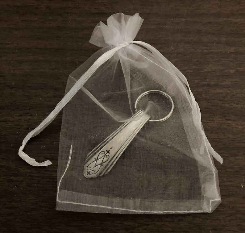 May include: A silver spoon keychain with a decorative design, packaged in a white organza bag.