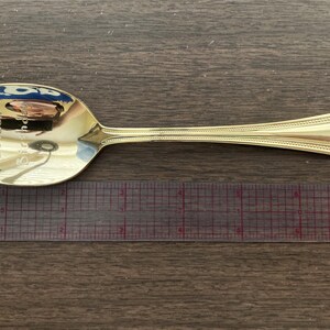 May include: A gold-colored spoon with the inscription "Happy Golden Birthday". The spoon is placed next to a ruler. The spoon has a decorative handle and a reflective surface.
