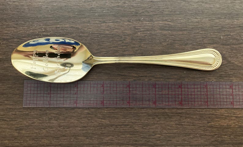 May include: A gold-colored spoon with the inscription "Happy Golden Birthday". The spoon is placed next to a ruler. The spoon has a decorative handle and a reflective surface.