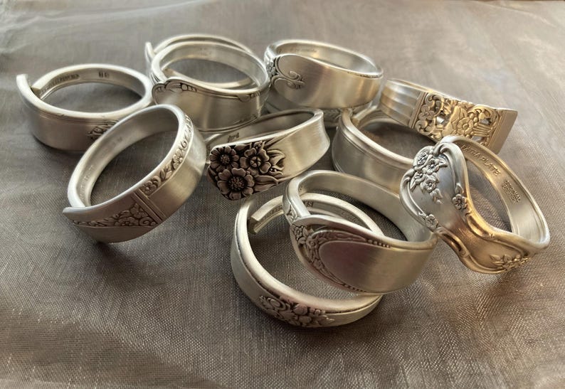 Puede incluir: Una colecci&oacute;n de nueve anillos de cuchara de plata, cada uno con un dise&ntilde;o &uacute;nico. Algunos anillos presentan patrones florales, mientras que otros tienen un dise&ntilde;o m&aacute;s simple. Todos los anillos est&aacute;n hechos de cubiertos reciclados.