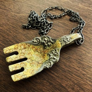 May include: A gold-toned fork pendant necklace with a silver chain. The fork has a marbled design and floral accents.