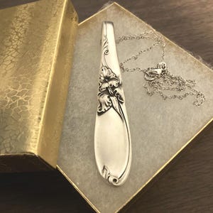 Puede incluir: Un collar con colgante de cuchara plateada con detalles florales, presentado en una caja de regalo dorada texturizada. El colgante de cuchara est&aacute; unido a una delicada cadena plateada. El collar se muestra sobre una superficie blanca texturizada.