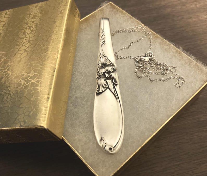Puede incluir: Un collar con colgante de cuchara plateada con detalles florales, presentado en una caja de regalo dorada texturizada. El colgante de cuchara est&aacute; unido a una delicada cadena plateada. El collar se muestra sobre una superficie blanca texturizada.
