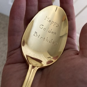 May include: A gold-colored spoon with the words "Happy Golden Birthday" engraved on the bowl. The spoon has a decorative handle and is held in a hand.